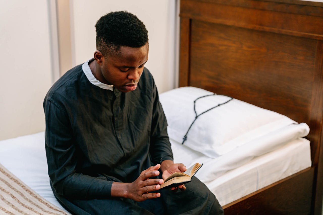 Seminarian reading a book on faith, seated on a bed in a peaceful room.